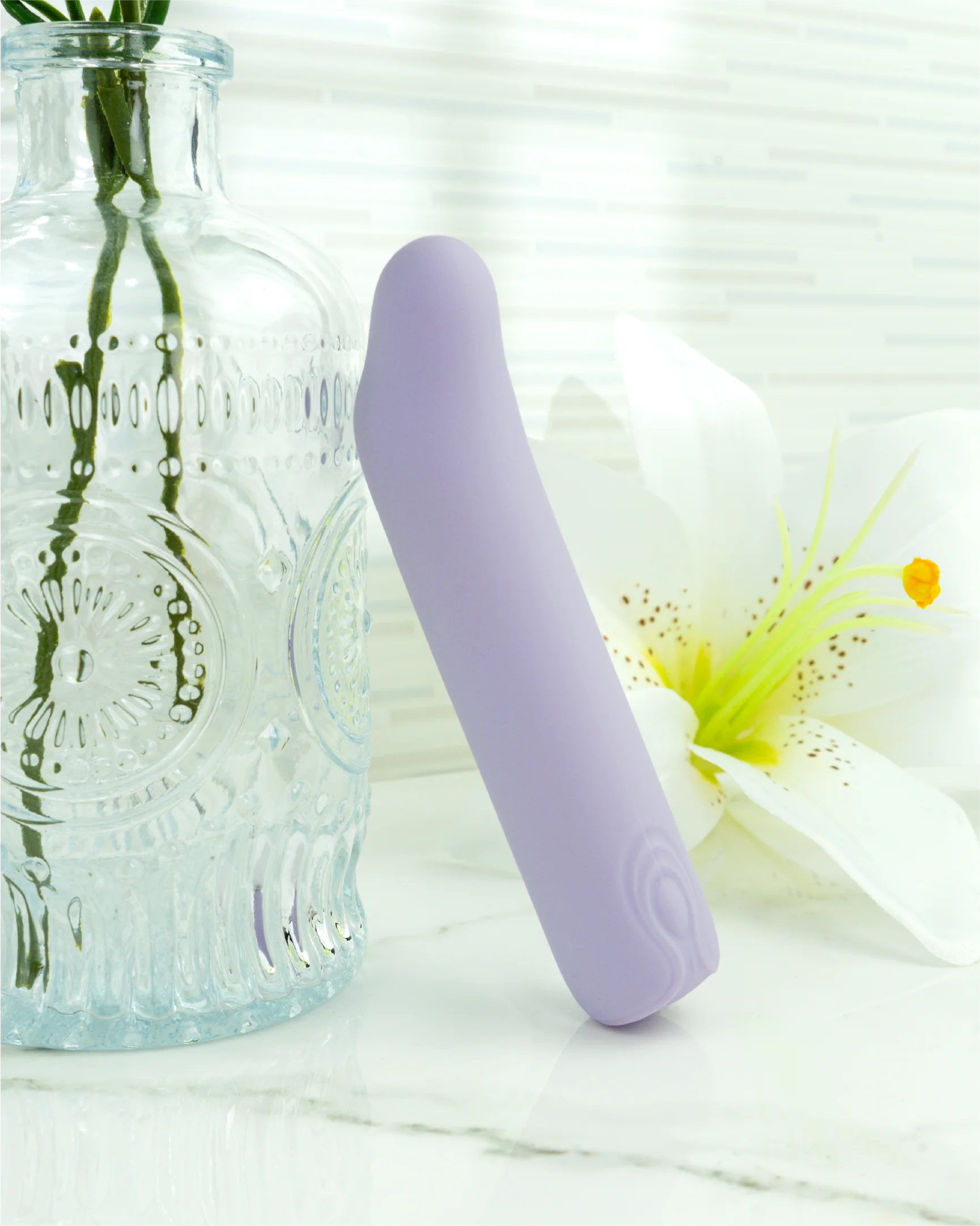 Pastel purple Blush & Bloom Iris curved mini G-spot vibrator beside glass bottle and flower on marble surface