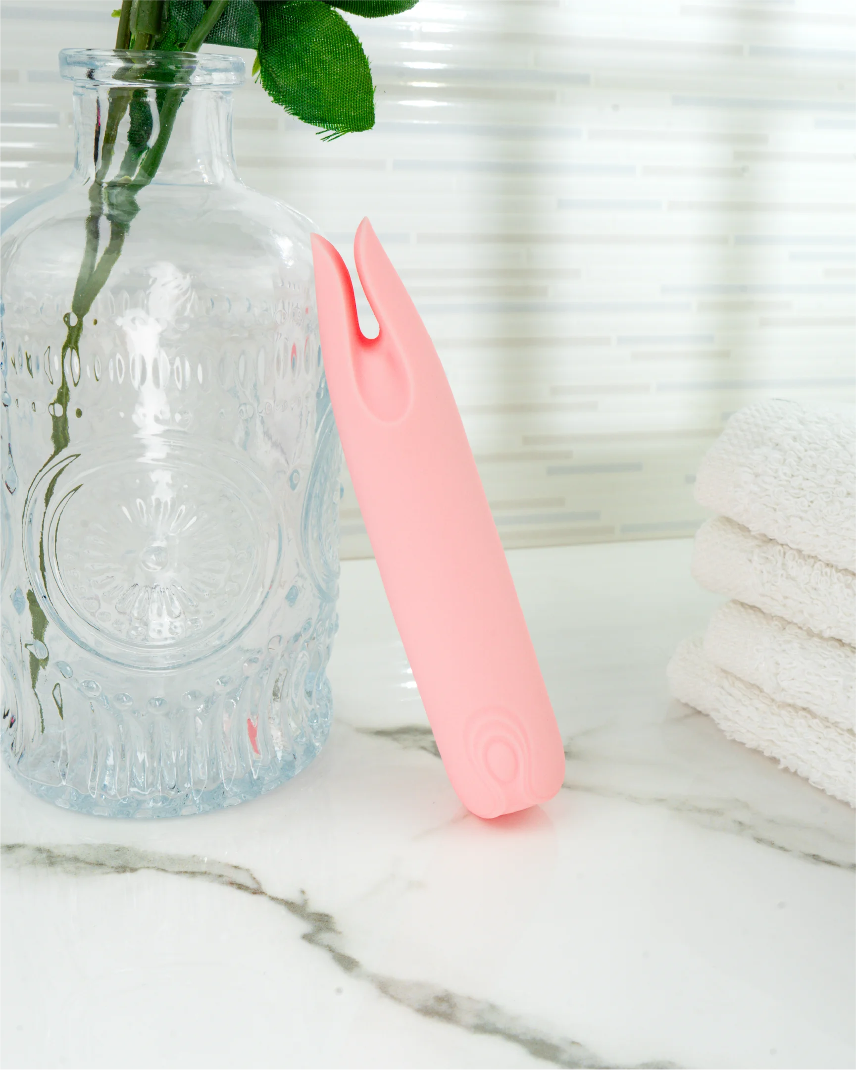 Pastel pink Petunia mini clitoral vibrator next to glass bottle on marble surface, waterproof silicone dual-tip design