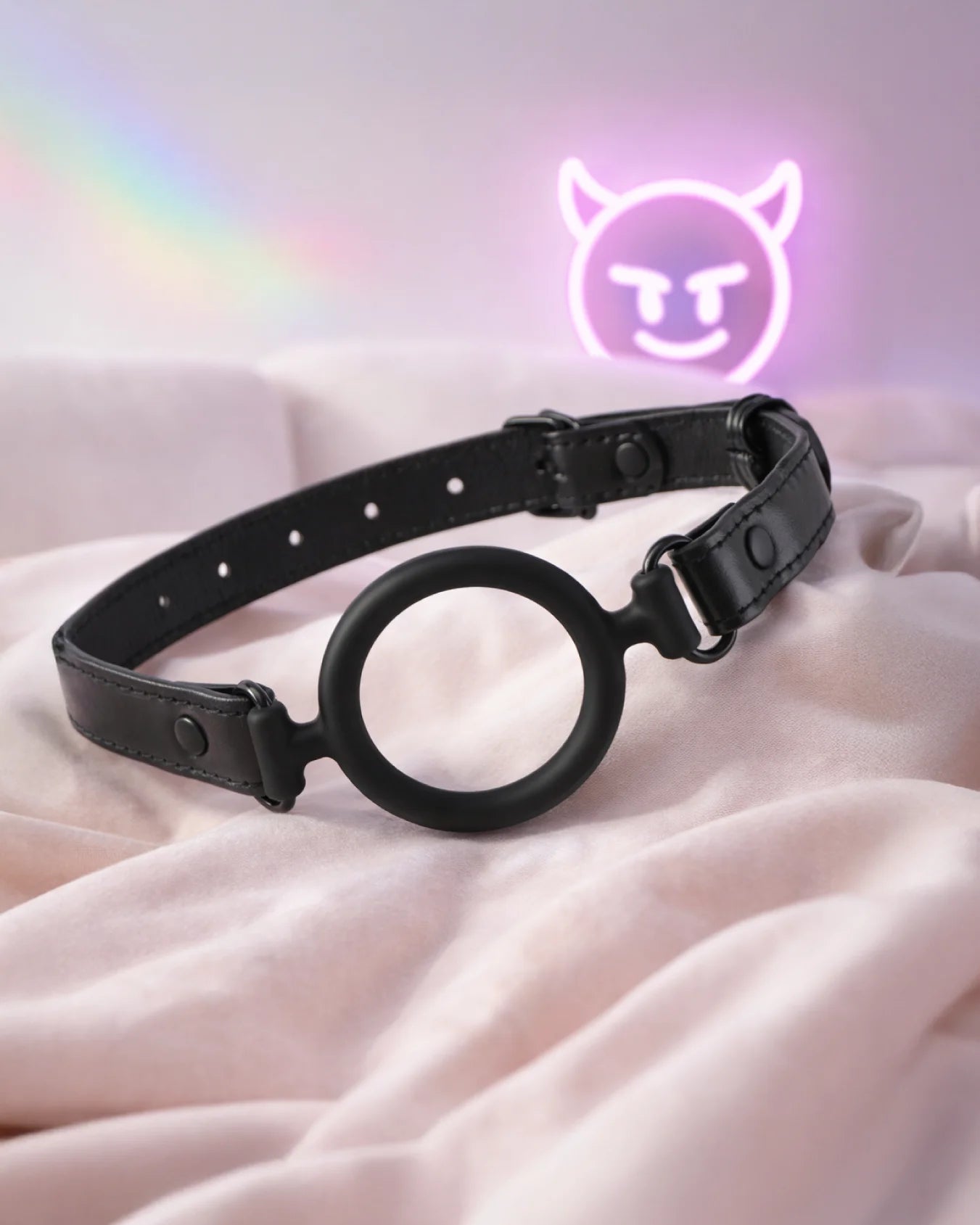 Black silicone O-ring gag on bed in soft bedroom setting with neon lighting, open-mouth gag for comfortable airflow