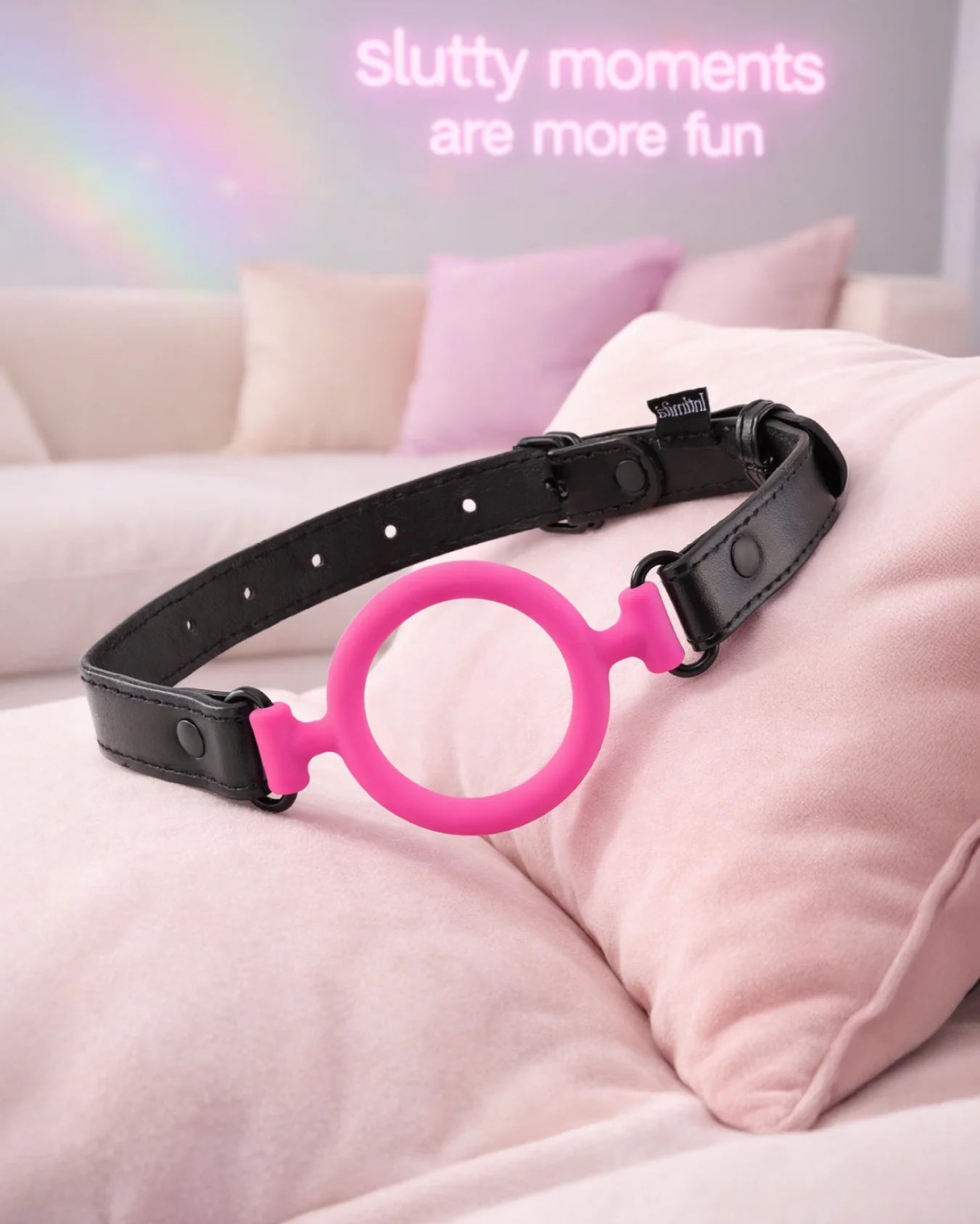 Pink silicone O-ring gag on pillow in soft bedroom scene, breathable open-mouth gag with adjustable strap design