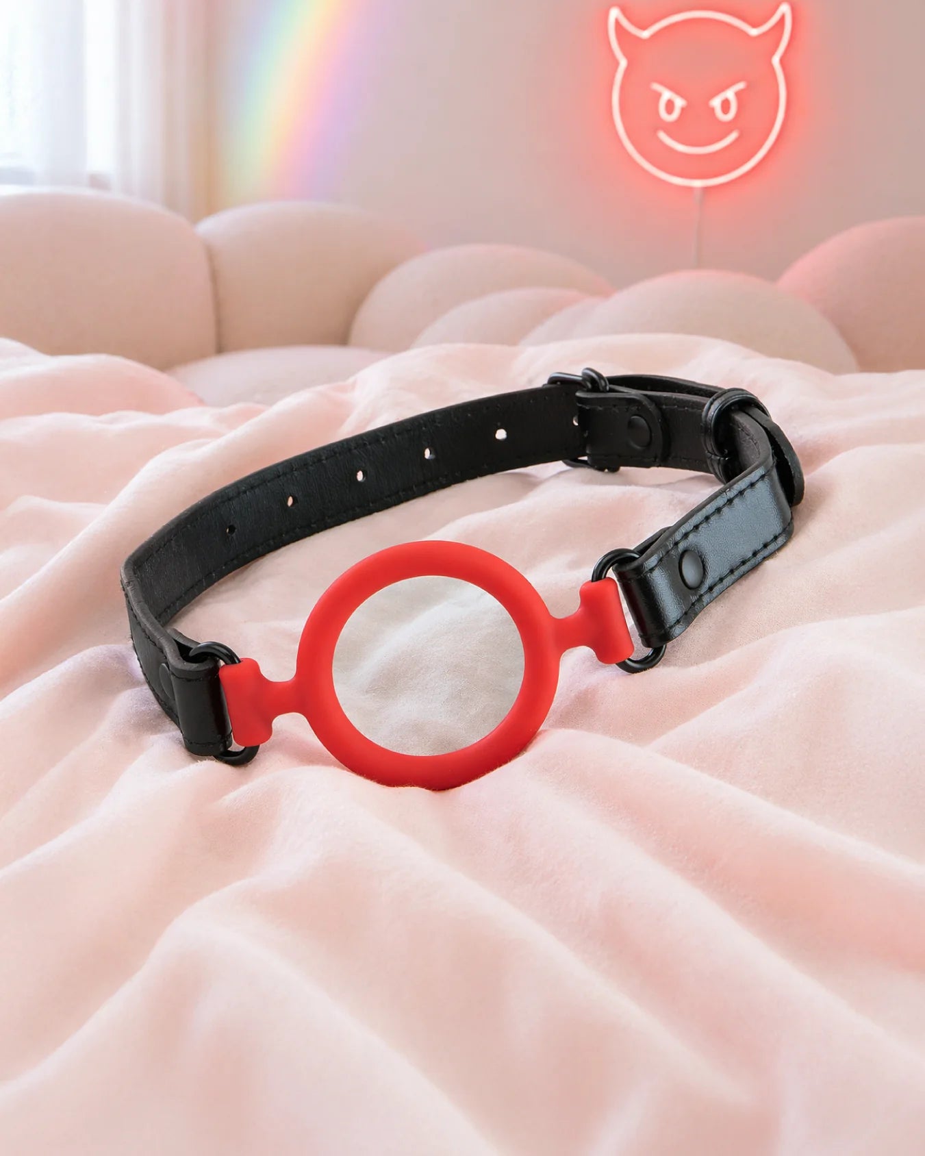 Red silicone O-ring gag resting on soft bed with neon background, open-mouth design for breathable mouth restraint