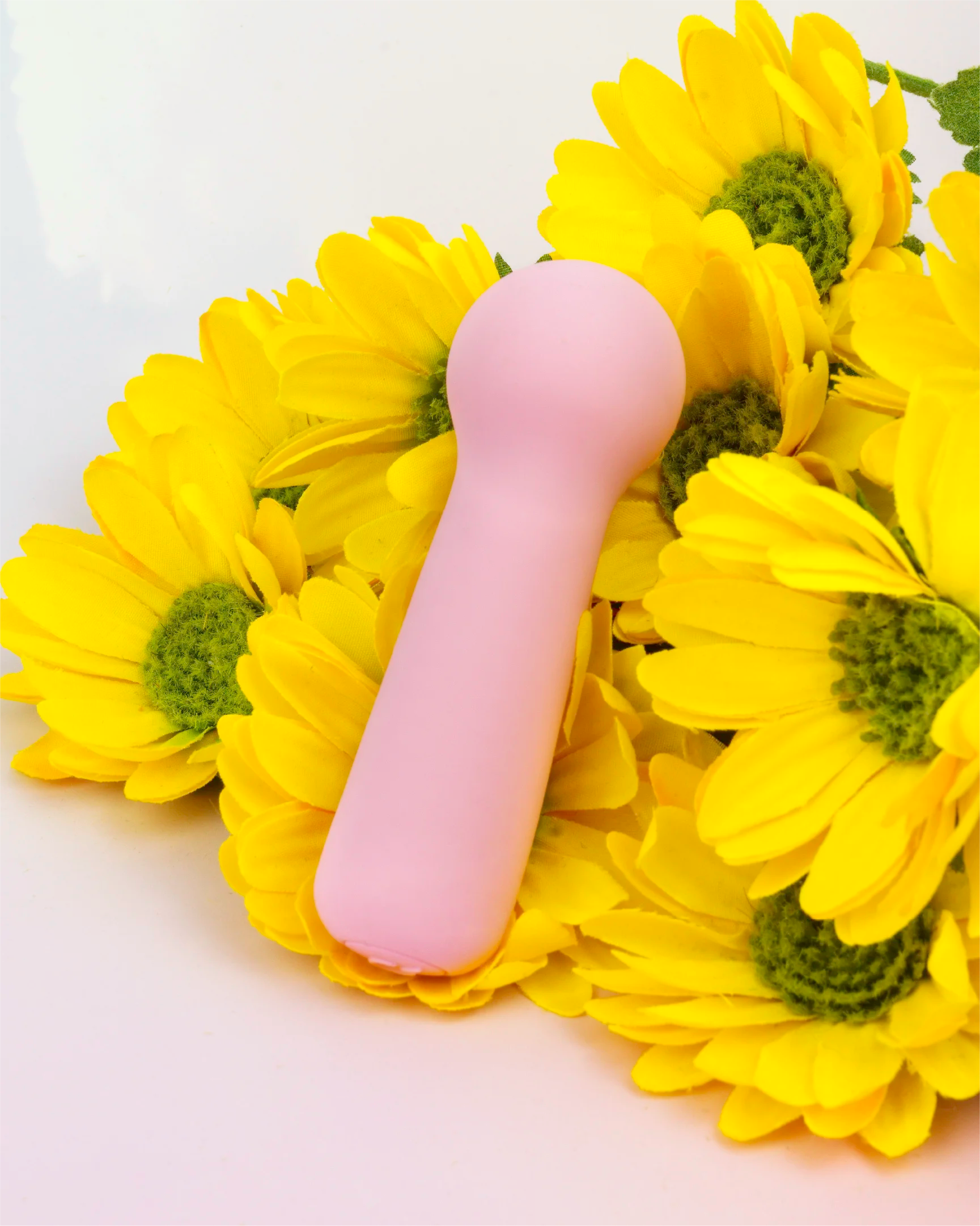 Pastel pink Joy Penny mini wand vibrator with rounded wand head displayed among yellow flowers, compact waterproof design with soft body-safe silicone